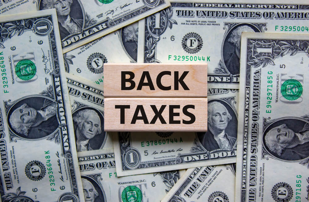 Wooden blocks displaying "BACK TAXES" on a background of U.S. one-dollar bills, illustrating the financial implications of unpaid taxes relevant to tax resolution services.