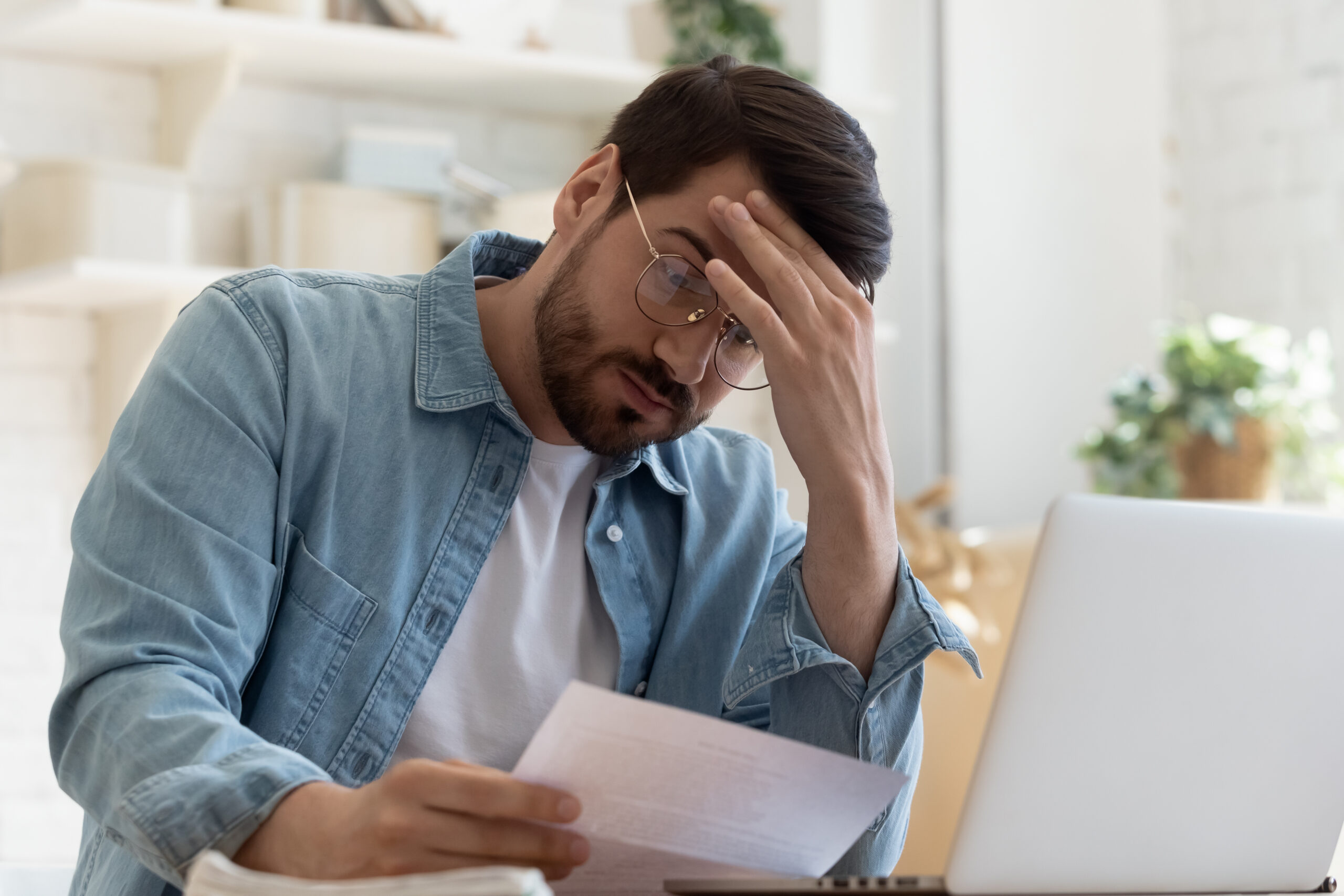 Concerned man reading IRS notice letter while sitting at a desk with a laptop, illustrating the anxiety of receiving tax-related communication.